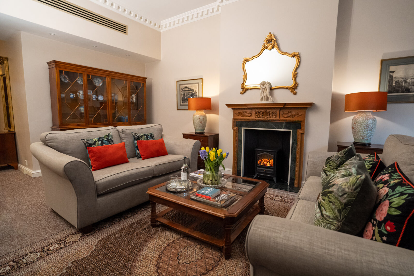 A period sitting room with fireplace, two grey settees either side, and a large square wood and glass coffee table.