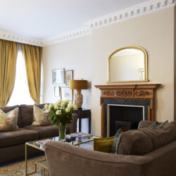 A period living room with feature fireplace, two brown settees, a coffee table holding a vase of white roses, and two period sash windows in the background, dressed with gold curtains.