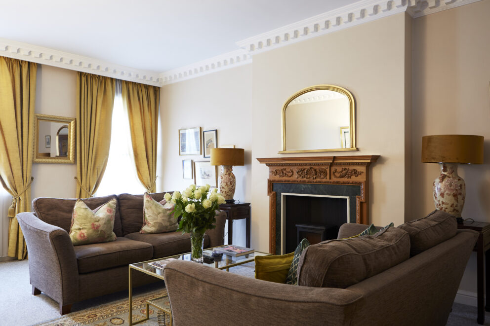 A period living room with feature fireplace, two brown settees, a coffee table holding a vase of white roses, and two period sash windows in the background, dressed with gold curtains.