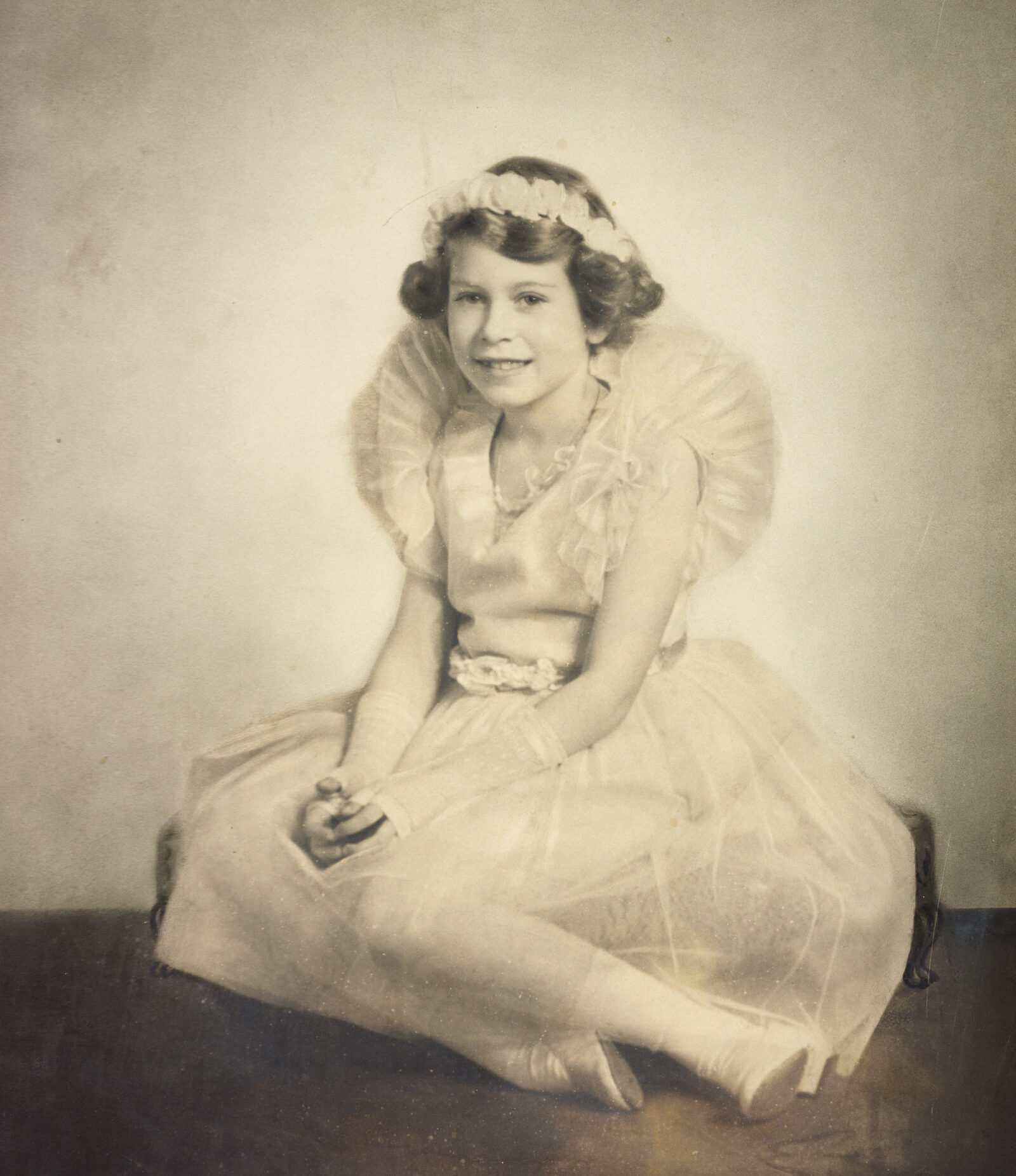 A sepia photo of Queen Elizabeth II as a child, seated in a dress with a headband.