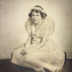 A sepia photo of Queen Elizabeth II as a child, seated in a dress with a headband.
