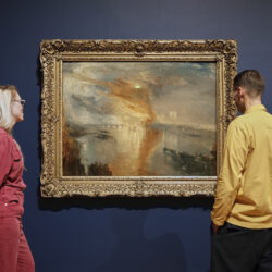 Two people stand either side of a Turner landscape painting, looking at the picture.