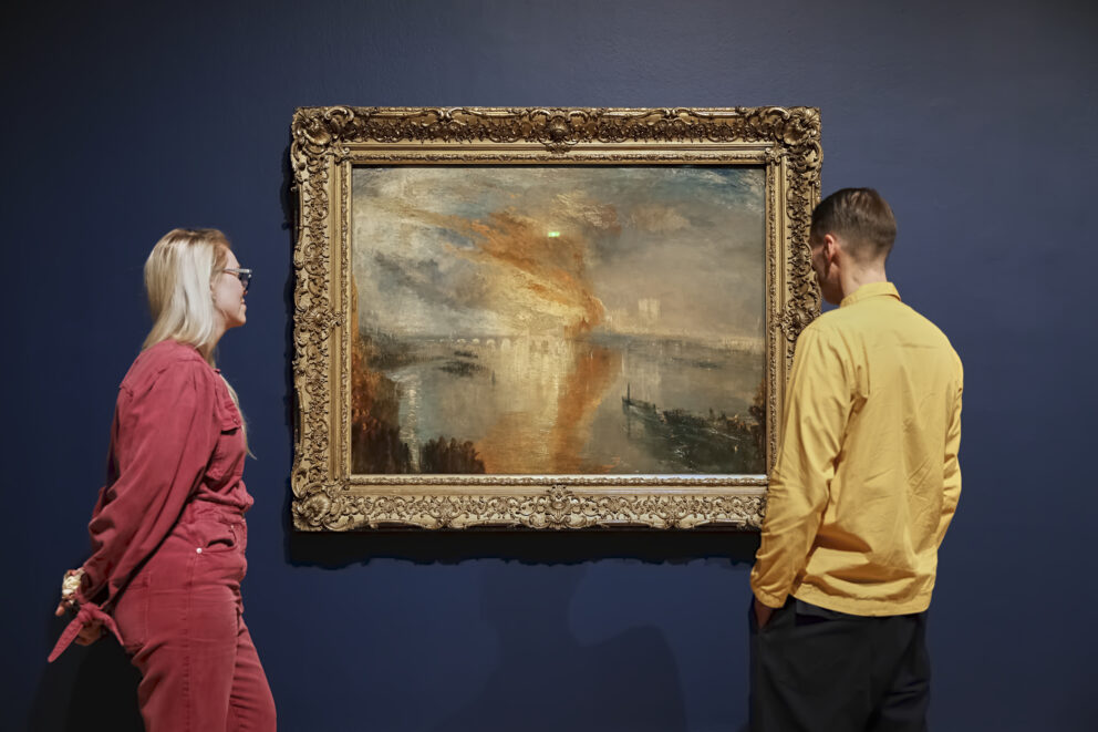 Two people stand either side of a Turner landscape painting, looking at the picture.
