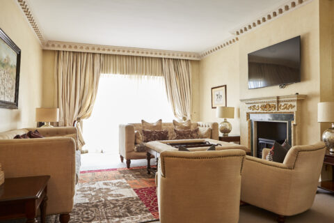Period sitting room with fireplace with tv above, cream settees and armchairs, a large square coffee table and doors leading out onto the balcony.