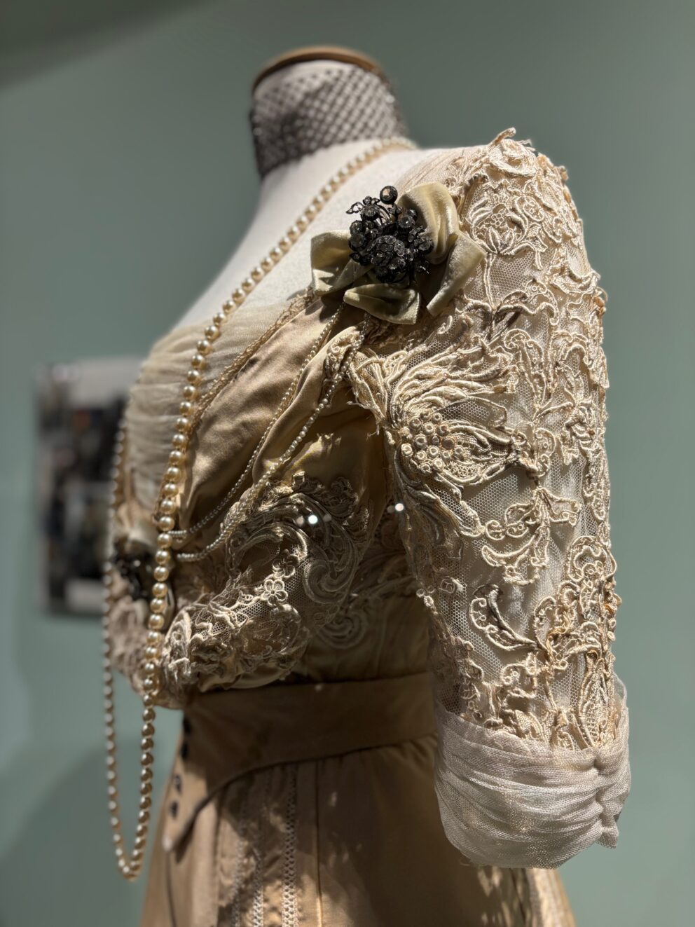 The cream lace and corsage detail of a period costume - the upper part of a dress, which is on a mannequin and accessorised with a pearl necklace.