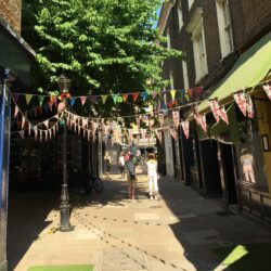 A pedestrianised alleyway with historic shops either side, bunting overhead and a tree in the background.