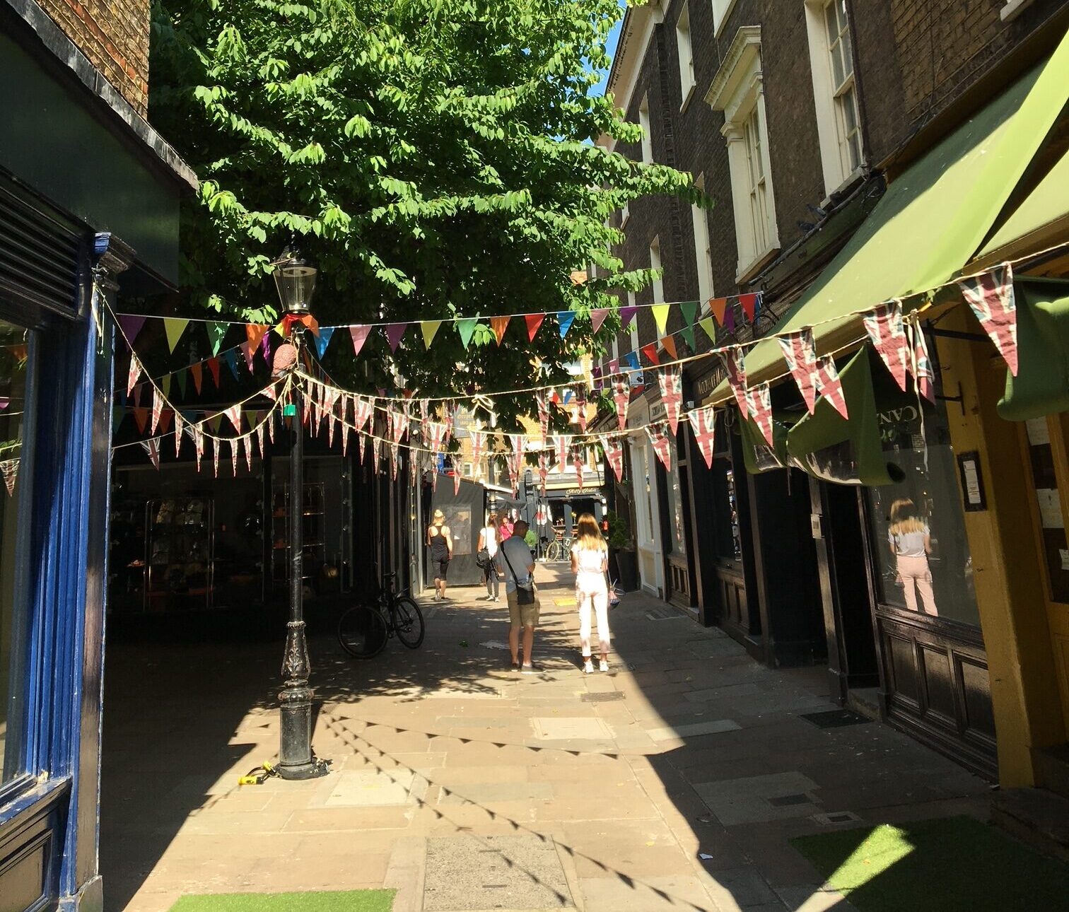 A pedestrianised alleyway with historic shops either side, bunting overhead and a tree in the background.