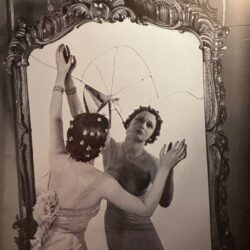 A sepia photograph of a woman in an evening dress, looking into an ornate but cracked mirror.