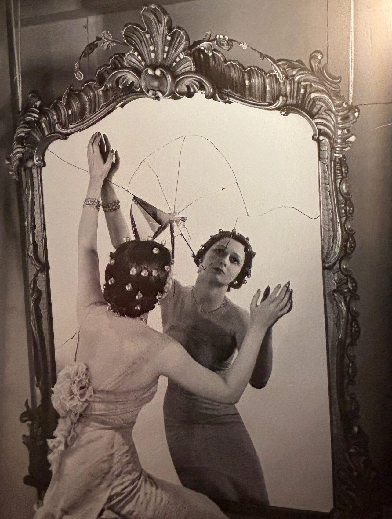 A sepia photograph of a woman in an evening dress, looking into an ornate but cracked mirror.