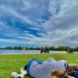 Two people lying on the grass in the sunshine by the Serpentine in Hyde Park.
