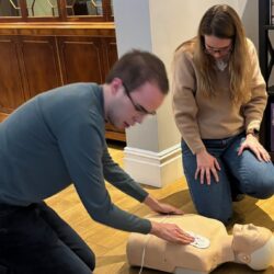 Two people kneeling on an apartment floor with a CPR mannequin which they are applying the defibrillator to.