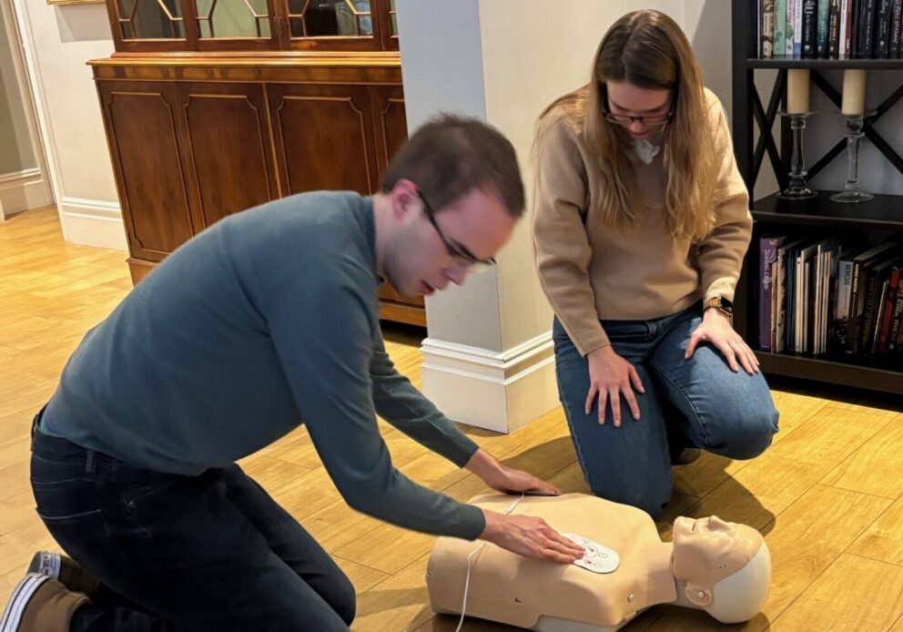 Two people kneeling on an apartment floor with a CPR mannequin which they are applying the defibrillator to.