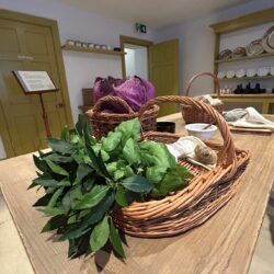 Handel's kitchen at Handel Hendrix House with vegetables in a wicker basket on a large wooden table in the centre of the room, and a large dresser in the background filled with plates on the racks.