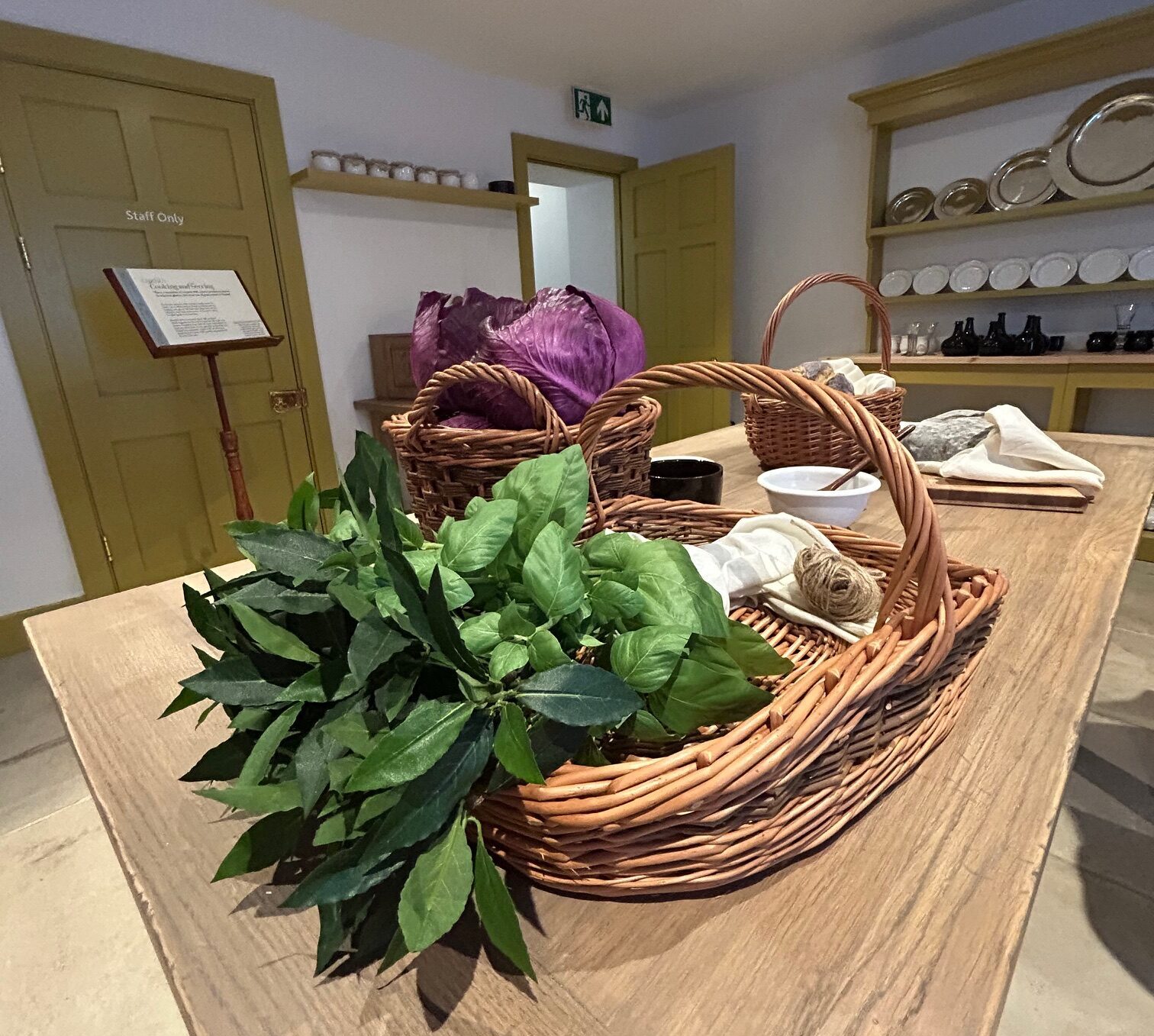 Handel's kitchen at Handel Hendrix House with vegetables in a wicker basket on a large wooden table in the centre of the room, and a large dresser in the background filled with plates on the racks.