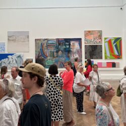 One of the galleries at the Royal Academy with the pale walls filled with artworks and a large group of visitors admiring the work.