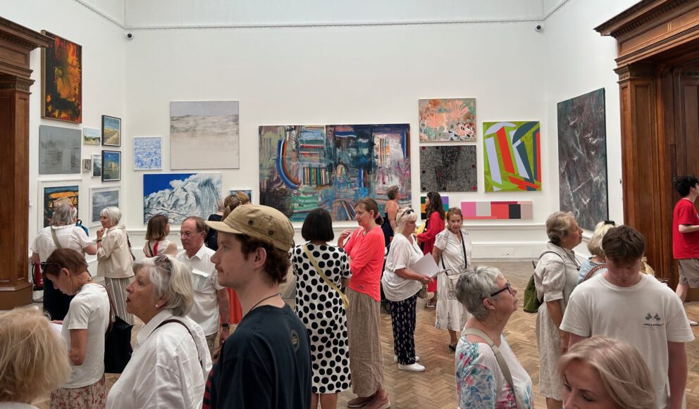 One of the galleries at the Royal Academy with the pale walls filled with artworks and a large group of visitors admiring the work.