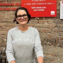 A woman wearing a grey jumper and dark trousers is standing at a shop desk, holding a dark garment.