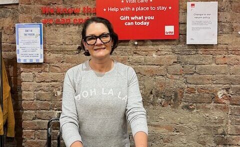 A woman wearing a grey jumper and dark trousers is standing at a shop desk, holding a dark garment.