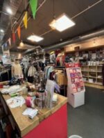 The interior of a Crisis shop, adorned with colourful bunting along the ceiling. There is a wooden and red painted desk, with garments on rails in the distance.