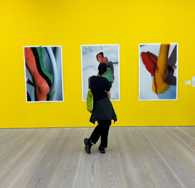 Woman in an art gallery, looking at three pictures hanging on a yellow wall.