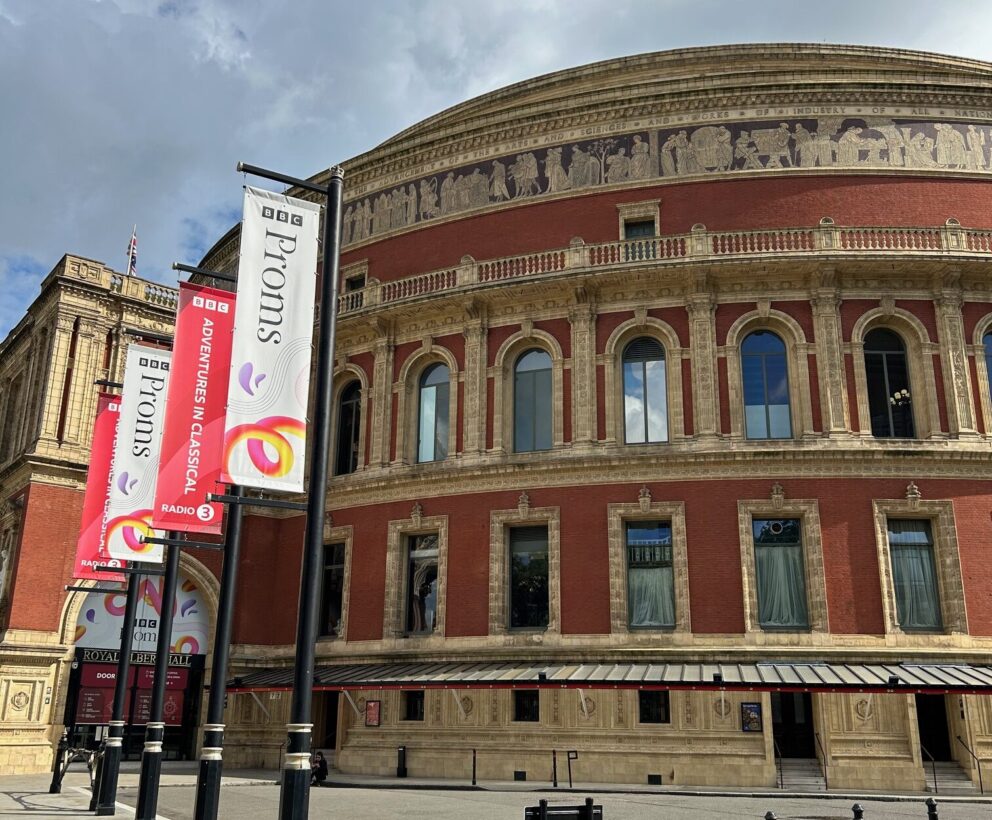 The exterior of the red brick circular Royal Albert Hall with vertical flags on black poles advertising The Proms.