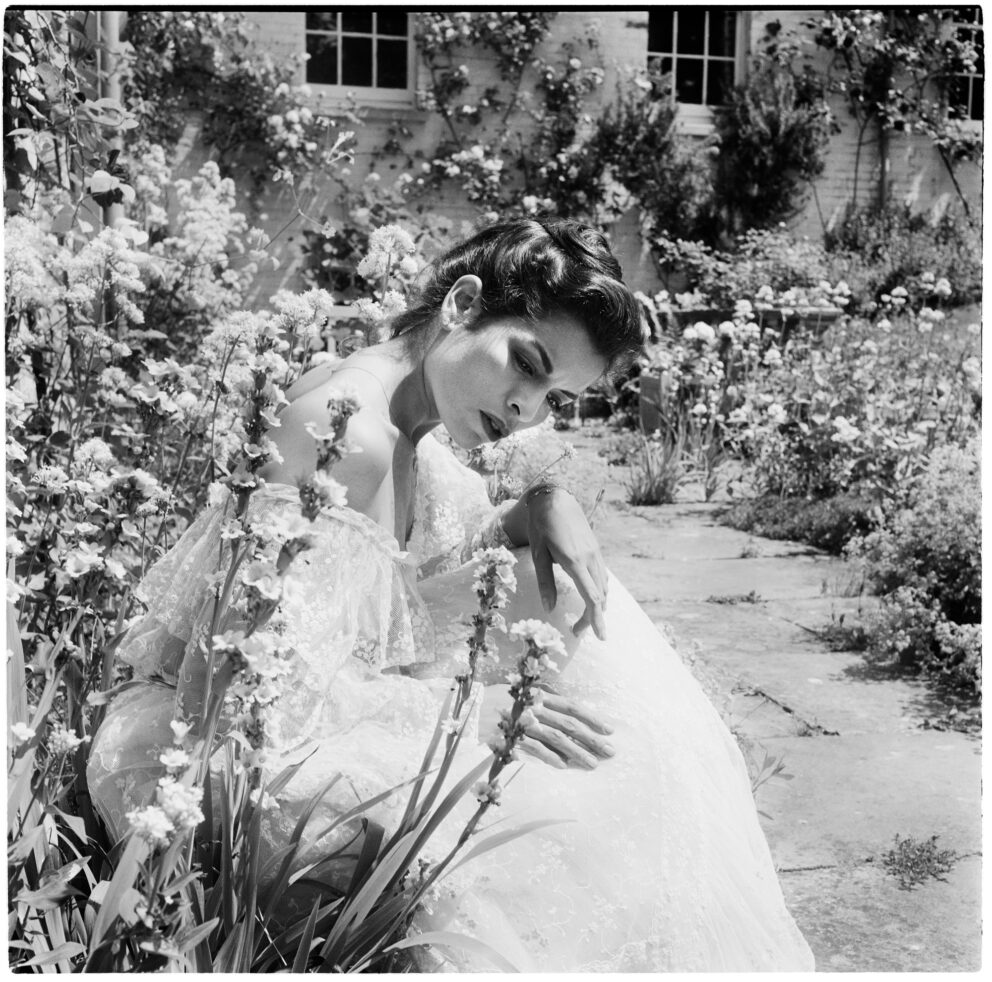 A black and white photo of Bianca Jagger in a long flowing dress, sitting among and looking at the flowers in a garden.