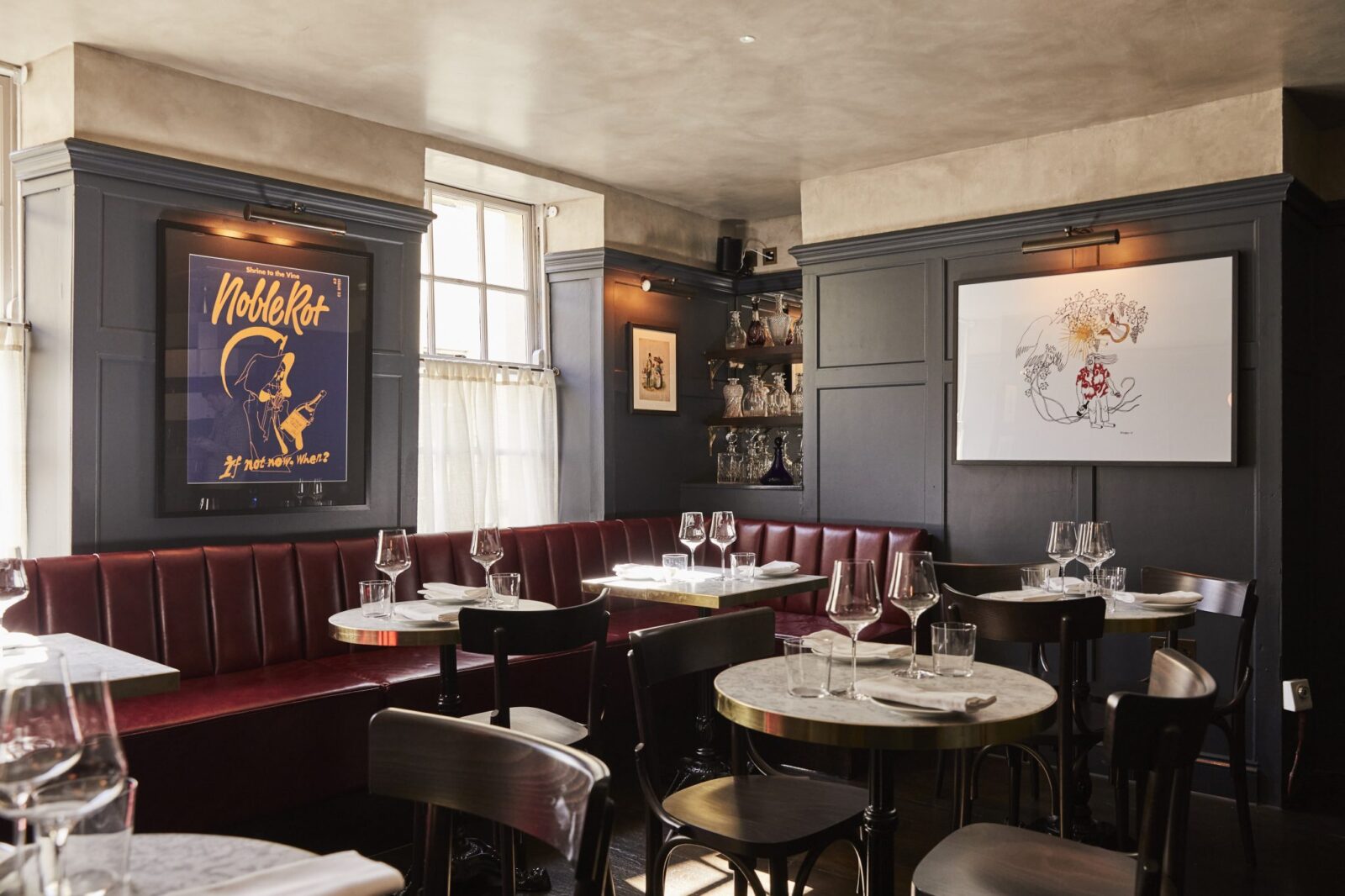 The interior of Noble Rot in Mayfair, with dark blue panelled walls, wooden tables and chairs, and plush red padded dining benches.