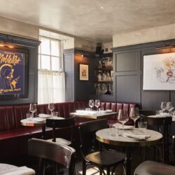 The interior of Noble Rot in Mayfair, with dark blue panelled walls, wooden tables and chairs, and plush red padded dining benches.