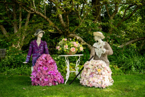 Two Downton Abbey characters created from floral displays, seated by a table in gardens.