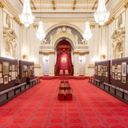 Two rows of display stands, exhibiting artworks, stand in the white, gold and red ballroom of Buckingham Palace.