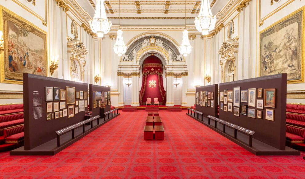 Two rows of display stands, exhibiting artworks, stand in the white, gold and red ballroom of Buckingham Palace.