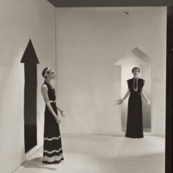 Two models, standing in a white room with arrows painted on walls and wearing dresses by Schiaparelli; Left in a dark crepe with bands of gold braids down the front, over satin pajamas; Right, in a black satin with bands of rippled gold ribbons.