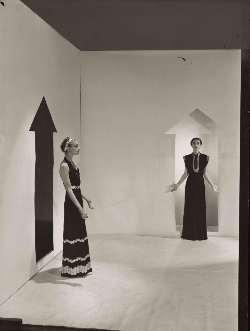 Two models, standing in a white room with arrows painted on walls and wearing dresses by Schiaparelli; Left in a dark crepe with bands of gold braids down the front, over satin pajamas; Right, in a black satin with bands of rippled gold ribbons.