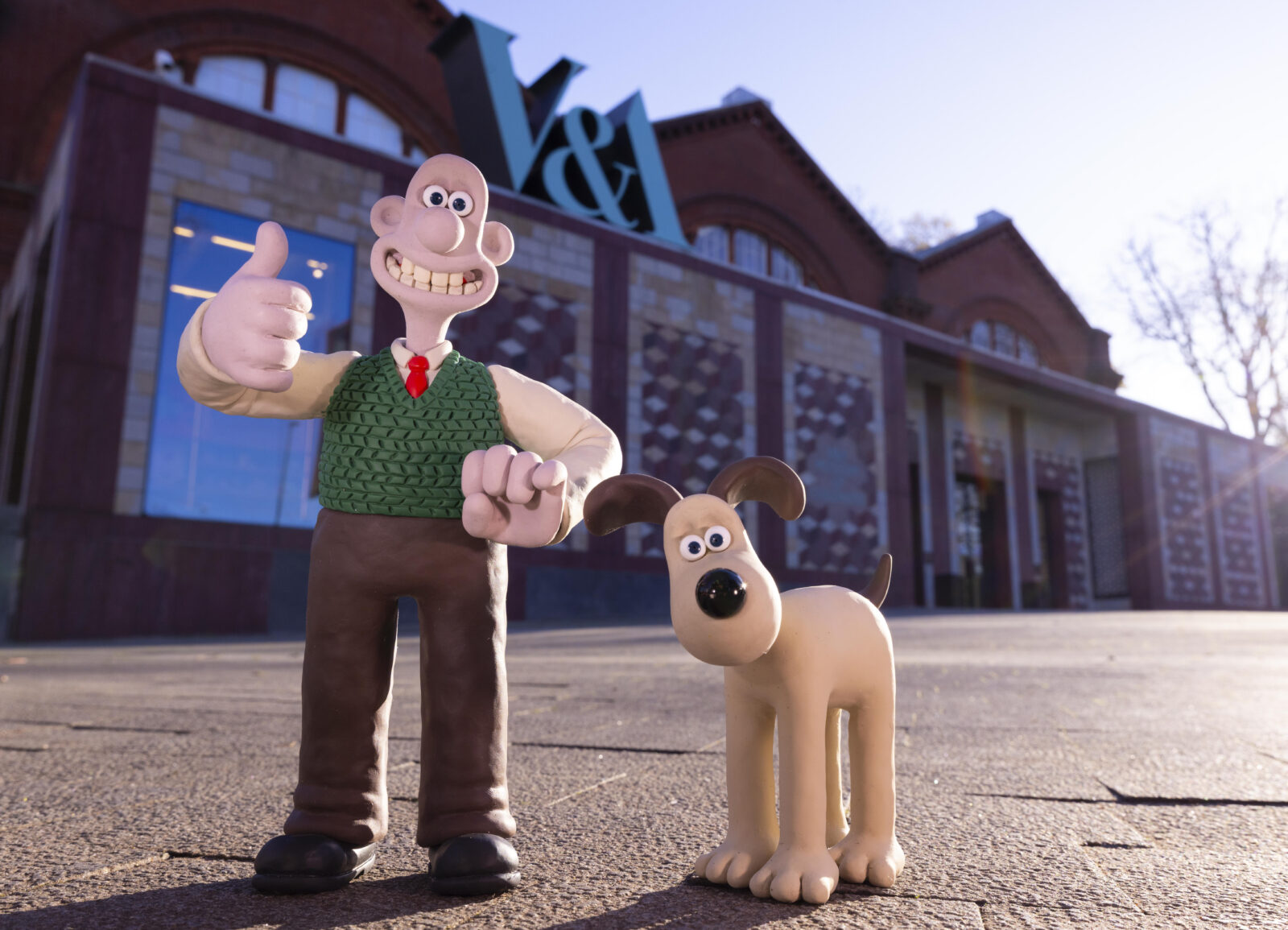 Wallace, wearing brown trousers, white shirt, red tie and green tank top, standing with a thumb up and smile, alongside cream dog Gromit, who has brown ears. They are outside the Young V&A museum.