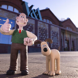 Wallace, wearing brown trousers, white shirt, red tie and green tank top, standing with a thumb up and smile, alongside cream dog Gromit, who has brown ears. They are outside the Young V&A museum.