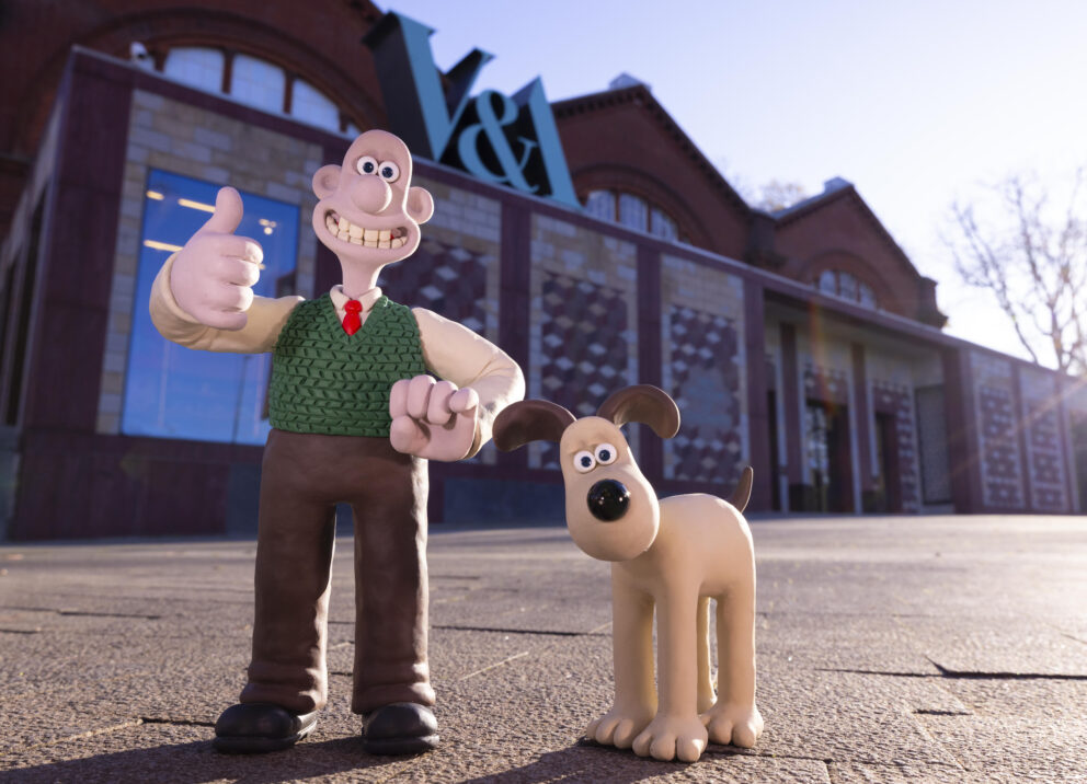 Wallace, wearing brown trousers, white shirt, red tie and green tank top, standing with a thumb up and smile, alongside cream dog Gromit, who has brown ears. They are outside the Young V&A museum.