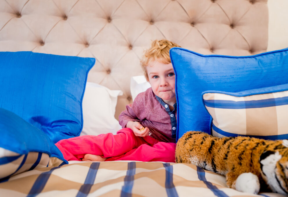 A young child is sitting on a bed, slightly hidden behind a cushion. His soft toy is on the bed beside him.