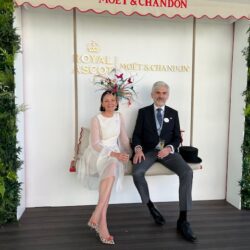 Our managing directors, Aude and Vladimir, seated on a white swing at Royal Ascot. Aude is wearing a white dress and multi-coloured headdress, Vladimir is in top hat and tails.