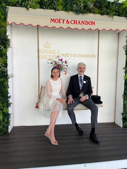 Our managing directors, Aude and Vladimir, seated on a white swing at Royal Ascot. Aude is wearing a white dress and multi-coloured headdress, Vladimir is in top hat and tails.
