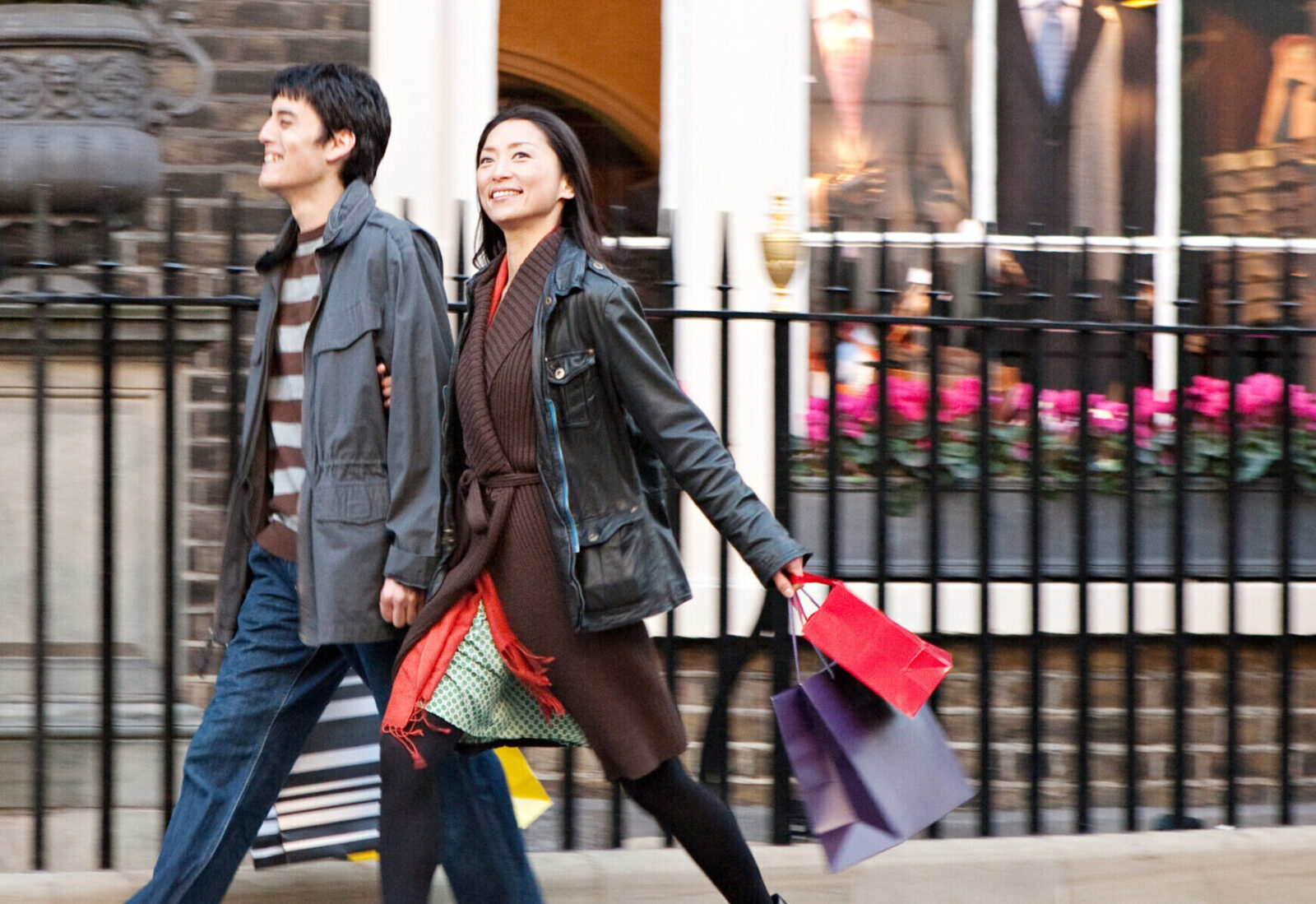 A man and a woman, dressed in cold weather clothing, walking along a period street, carrying paper boutique-style shopping bags.