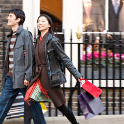 A man and a woman, dressed in cold weather clothing, walking along a period street, carrying paper boutique-style shopping bags.