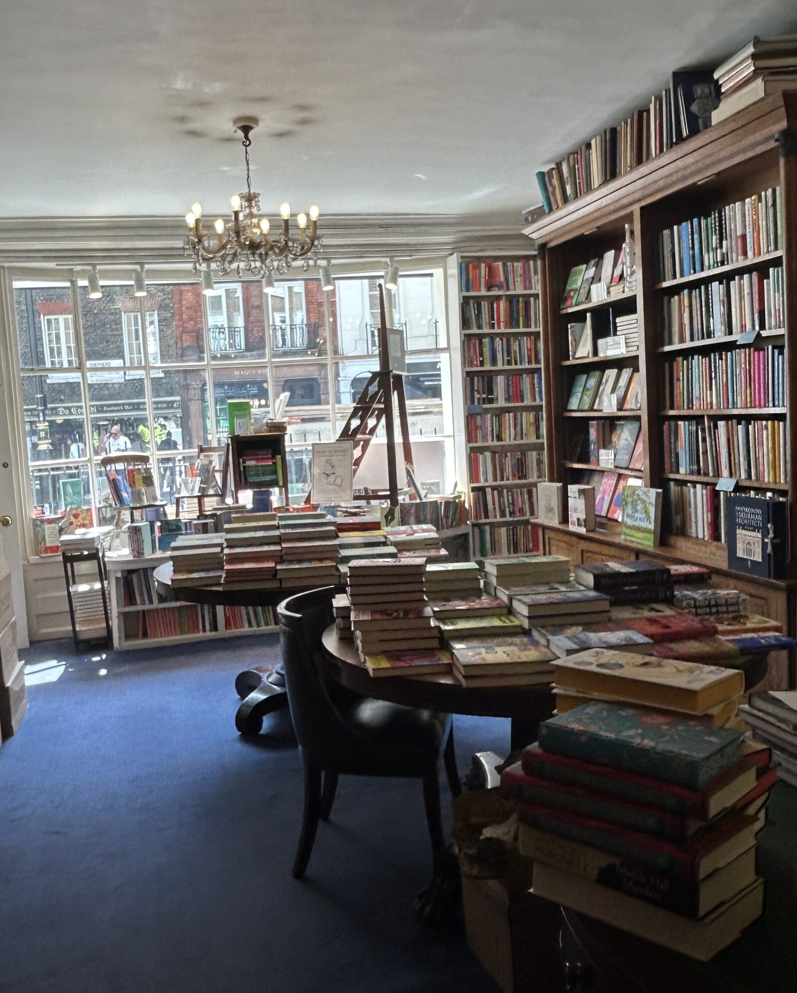 The interior of Heywood Hill bookshop with wooden bookshelves, table, chair and desk. The bookshelves are lined with books, there is a lit chandelier and a royal blue carpet.