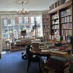 The interior of Heywood Hill bookshop with wooden bookshelves, table, chair and desk. The bookshelves are lined with books, there is a lit chandelier and a royal blue carpet.