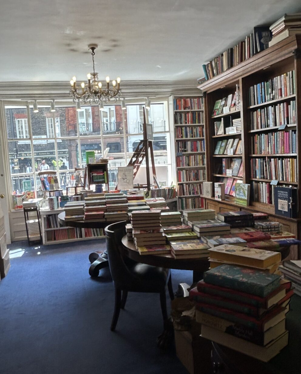The interior of Heywood Hill bookshop with wooden bookshelves, table, chair and desk. The bookshelves are lined with books, there is a lit chandelier and a royal blue carpet.