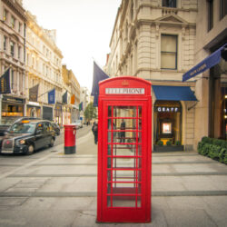 A photograph of a shopping street in Mayfair lined with designer boutiques. In the foreground there is a red telephone box and in the distance there is a red post box and a black London taxi.