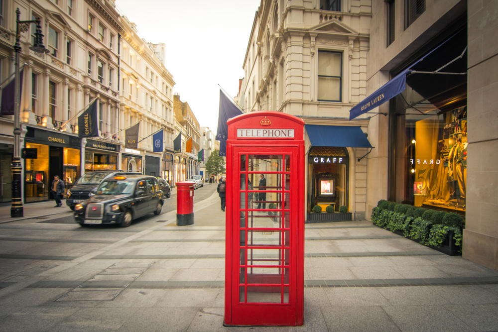 A photograph of a shopping street in Mayfair lined with designer boutiques. In the foreground there is a red telephone box and in the distance there is a red post box and a black London taxi.