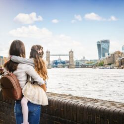 A woman is holding a child; they are both looking across the River Thames to Tower Bridge.