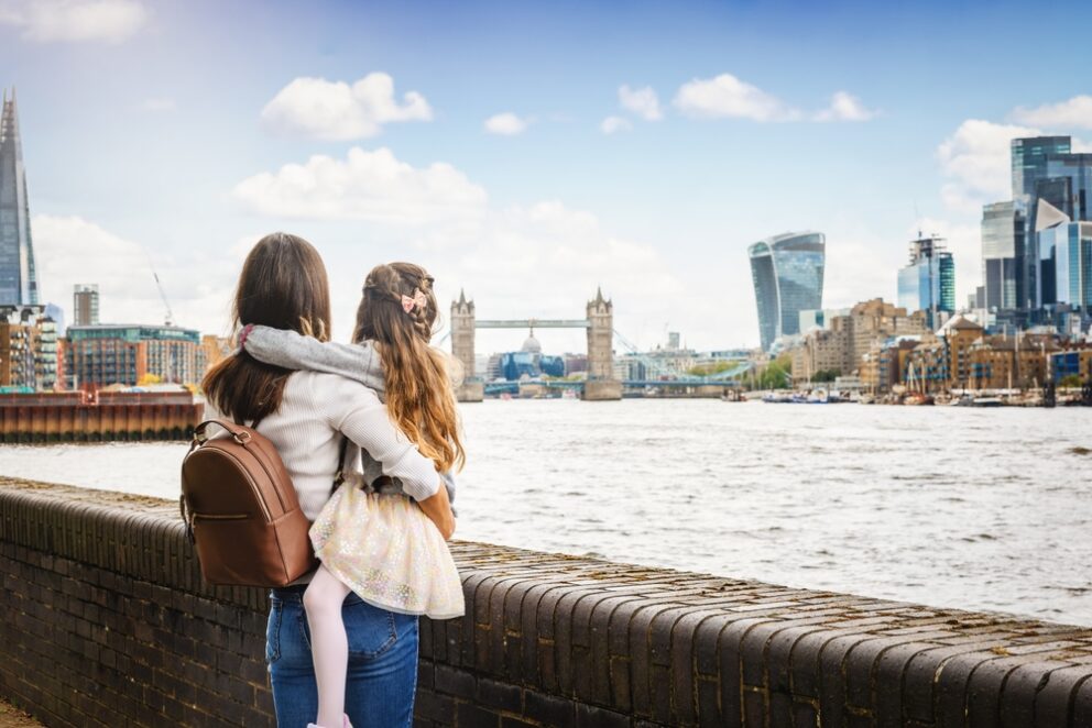 A woman is holding a child; they are both looking across the River Thames to Tower Bridge.