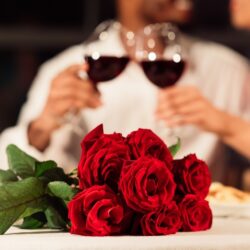A bouquet of red roses on a table in the foreground, with a couple drinking red wine behind.