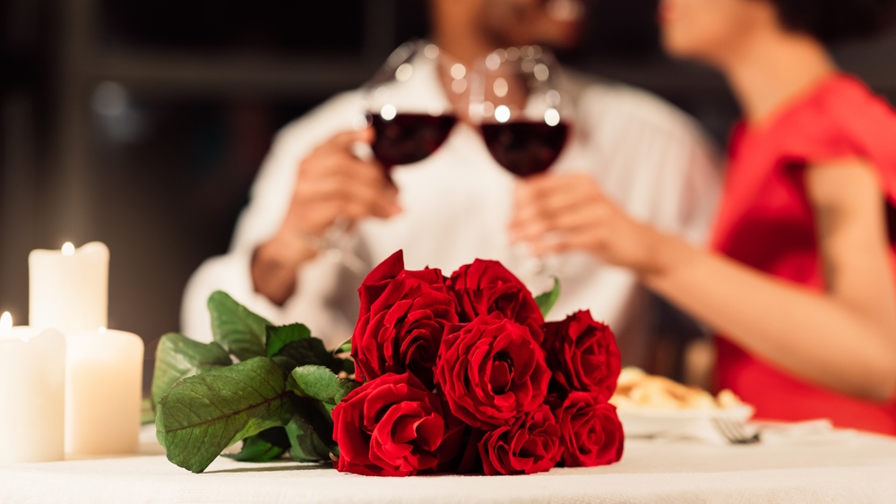 A bouquet of red roses on a table in the foreground, with a couple drinking red wine behind.